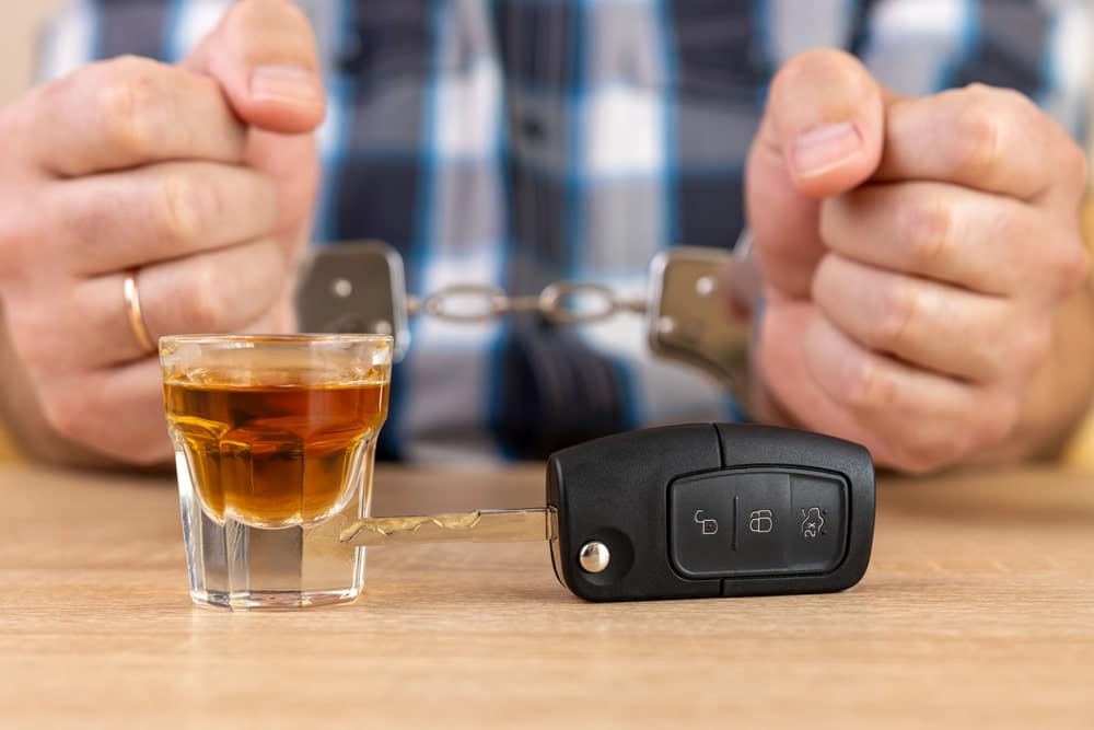 Image shows a person in handcuffs behind a shot glass and car key, concept of a DUI lawyer handling drunk driving arrest issues.