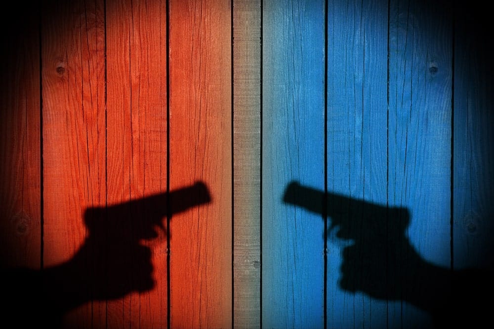 Image shows two shadowed hands pointing handguns toward each other against red and blue wooden panels, representing conflict and the need for a gun-weapon offenses lawyer.