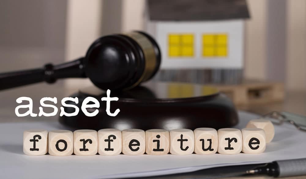 Image is of wooden blocks spelling asset forfeiture with a gavel and model house, concept of seizure of property lawyer handling forfeiture cases.